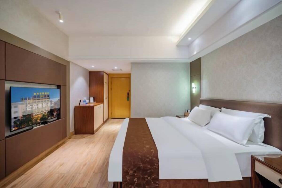A hotel room with a bed, a TV mounted on the wall, a wooden desk, and a door leading to another room.