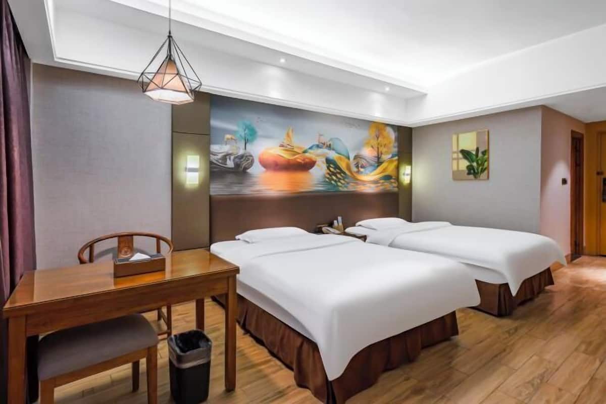 A hotel room with two beds, a wooden desk, a trash bin, a painting on the wall, and a ceiling light fixture.
