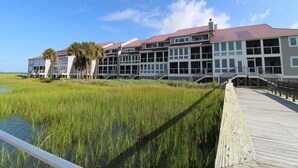 A waterfront hotel with multiple balconies, a wooden boardwalk, and a grassy area with tall grass.