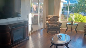 A living room with a flat-screen TV, wooden flooring, a ceiling fan, a patterned armchair, a glass coffee table, and a view of a garden through large windows. - Suncity west Golf course community (Sun City West)