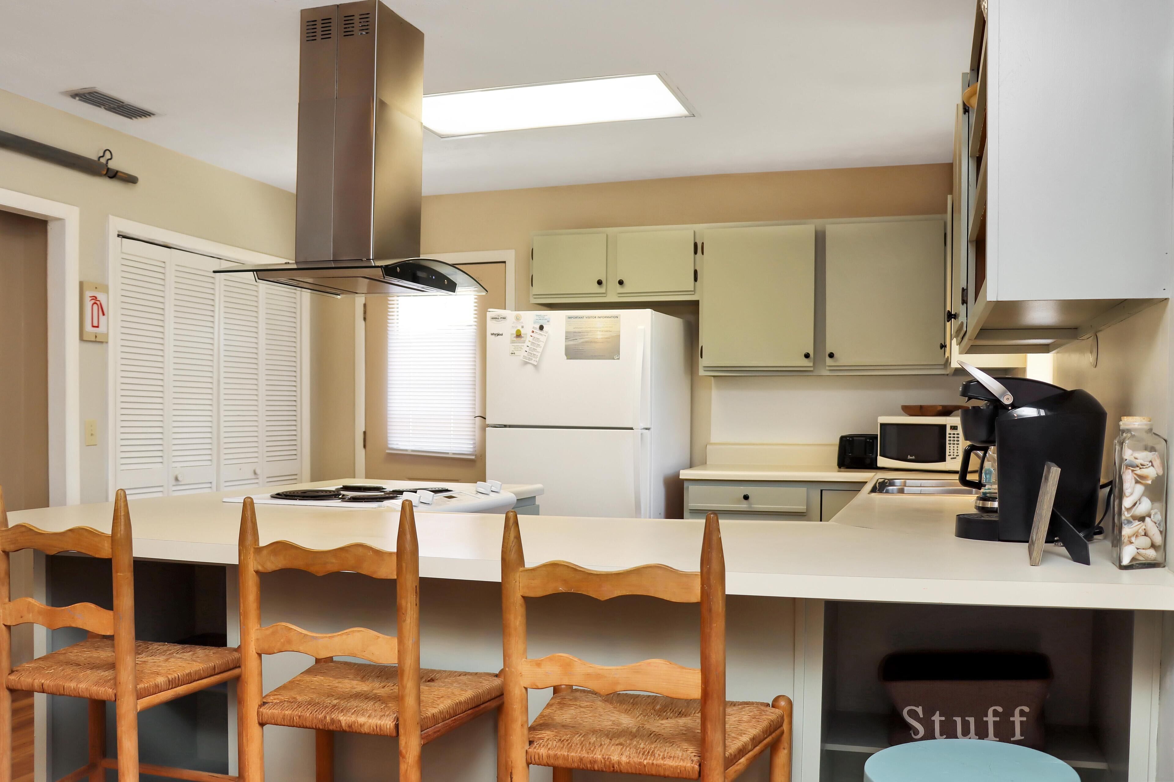 A compact kitchen with a white countertop, a built-in microwave, a coffee maker, and wooden chairs with woven seats.