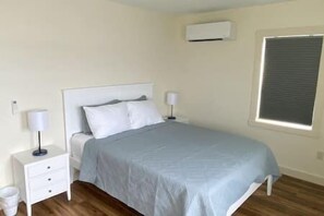 A bedroom with a bed, bedside tables, a lamp, an air conditioning unit, and a window with blinds. - #48 Isle View - Sleeps 6, Sunsets & Beach (Alburgh)