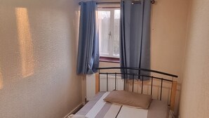 Ein kleines Zimmer mit einem Einzelbett, einem Metallgestell, einer Deckenleuchte, einem Fenster mit Vorhängen und einem Heizkörper.