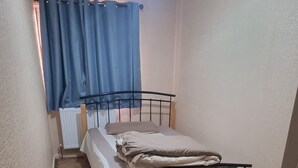 Ein kleiner Raum mit Teppichboden, einem Einzelbett, einem Heizkörper und einem Fenster mit blauen Vorhängen.