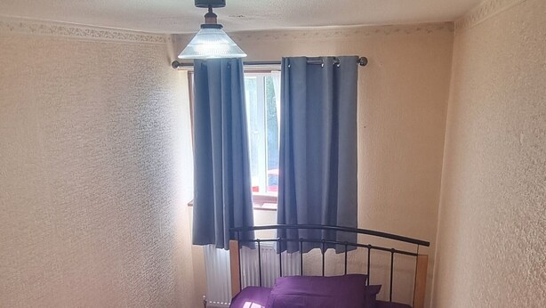 Ein kleines Zimmer mit einem Einzelbett, einer violetten Tagesdecke, einem weißen Kopfkissen und einem kleinen Tisch.