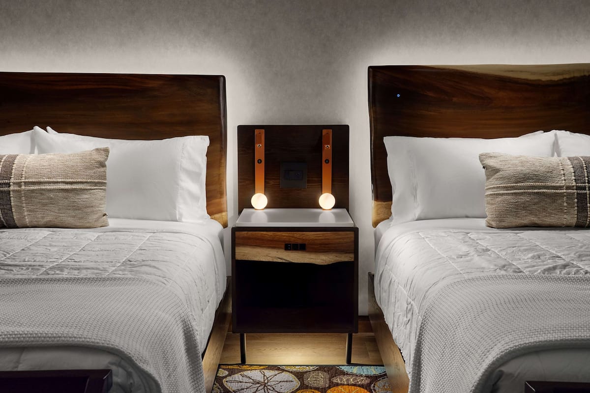 A hotel room with two beds, each with a wooden headboard and white bedding. There is a nightstand between the beds with two illuminated bulbs.