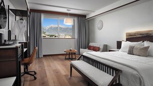 Una camera d'hotel con un letto, una scrivania, una sedia, un tavolino, una panca, una finestra vista montagna e una sveglia a parete.