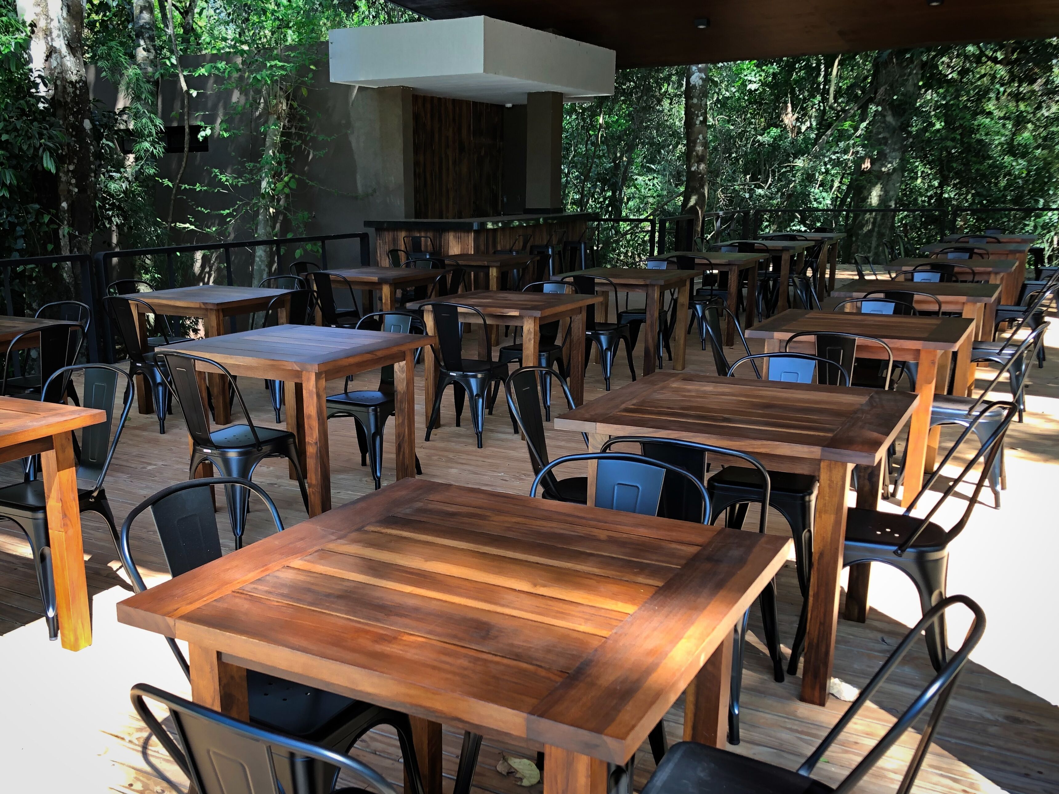 Un área de comedor al aire libre con varias mesas de madera y sillas de metal, rodeada de árboles.