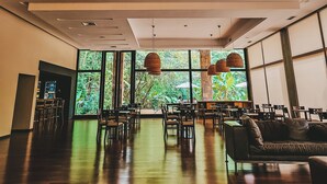 A spacious indoor area with multiple hanging pendant lights, large windows offering a view of greenery outside, and a seating area with tables and chairs. - Hotel Tre (Puerto Iguazú)