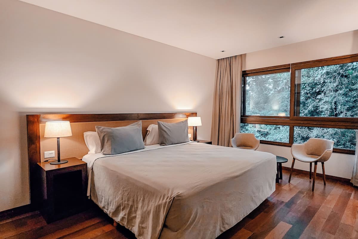 A modern hotel room with a large bed, two bedside tables with lamps, a small round table, and a chair. The room has wooden flooring and a large window with a view of trees.