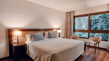 A modern hotel room with a large bed, two bedside tables with lamps, a small round table, and a chair. The room has wooden flooring and a large window with a view of trees.