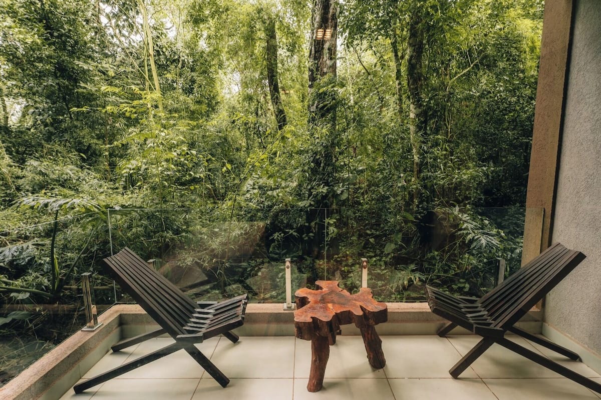 A modern outdoor seating area with two chairs and a wooden table, overlooking a lush, green forest.