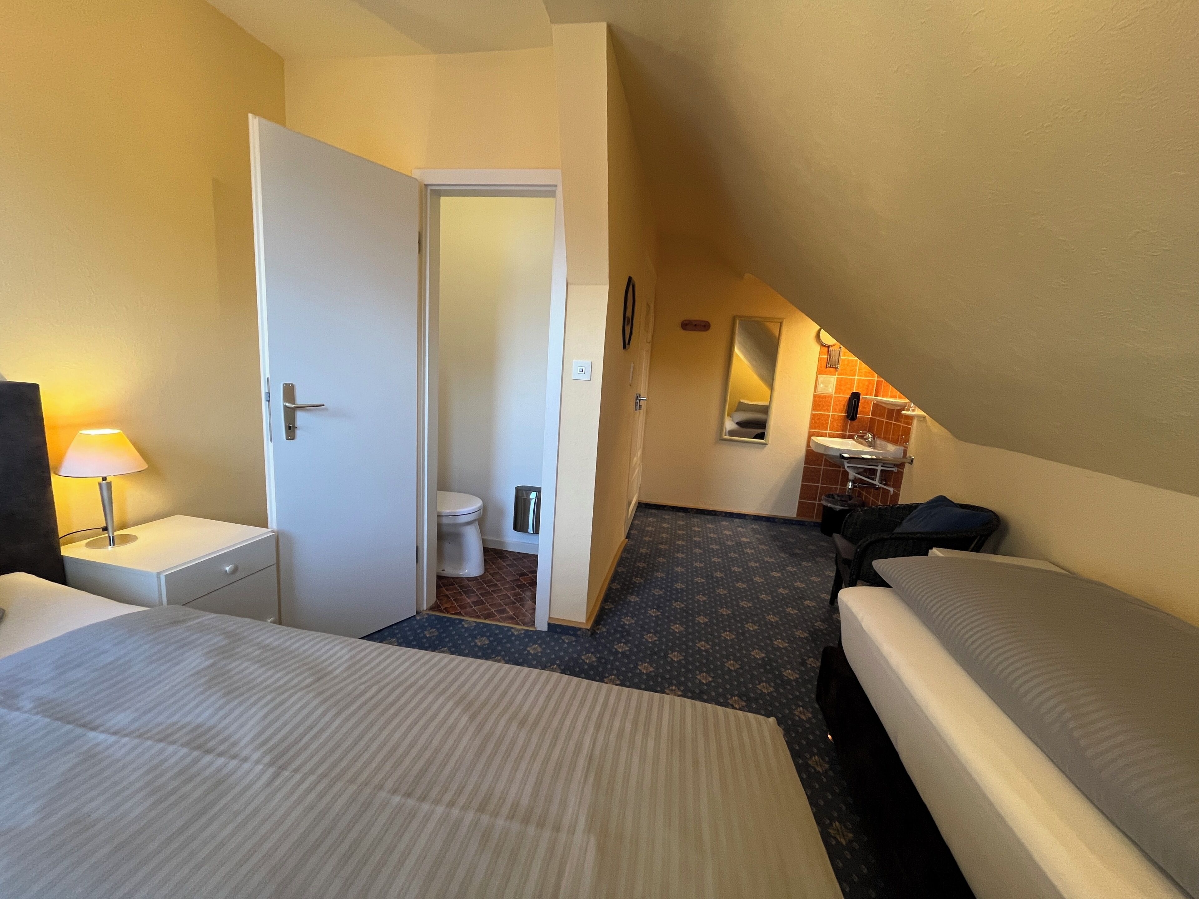 A hotel room with two beds, a nightstand with a lamp, a bathroom with a toilet and sink, and a small mirror.
