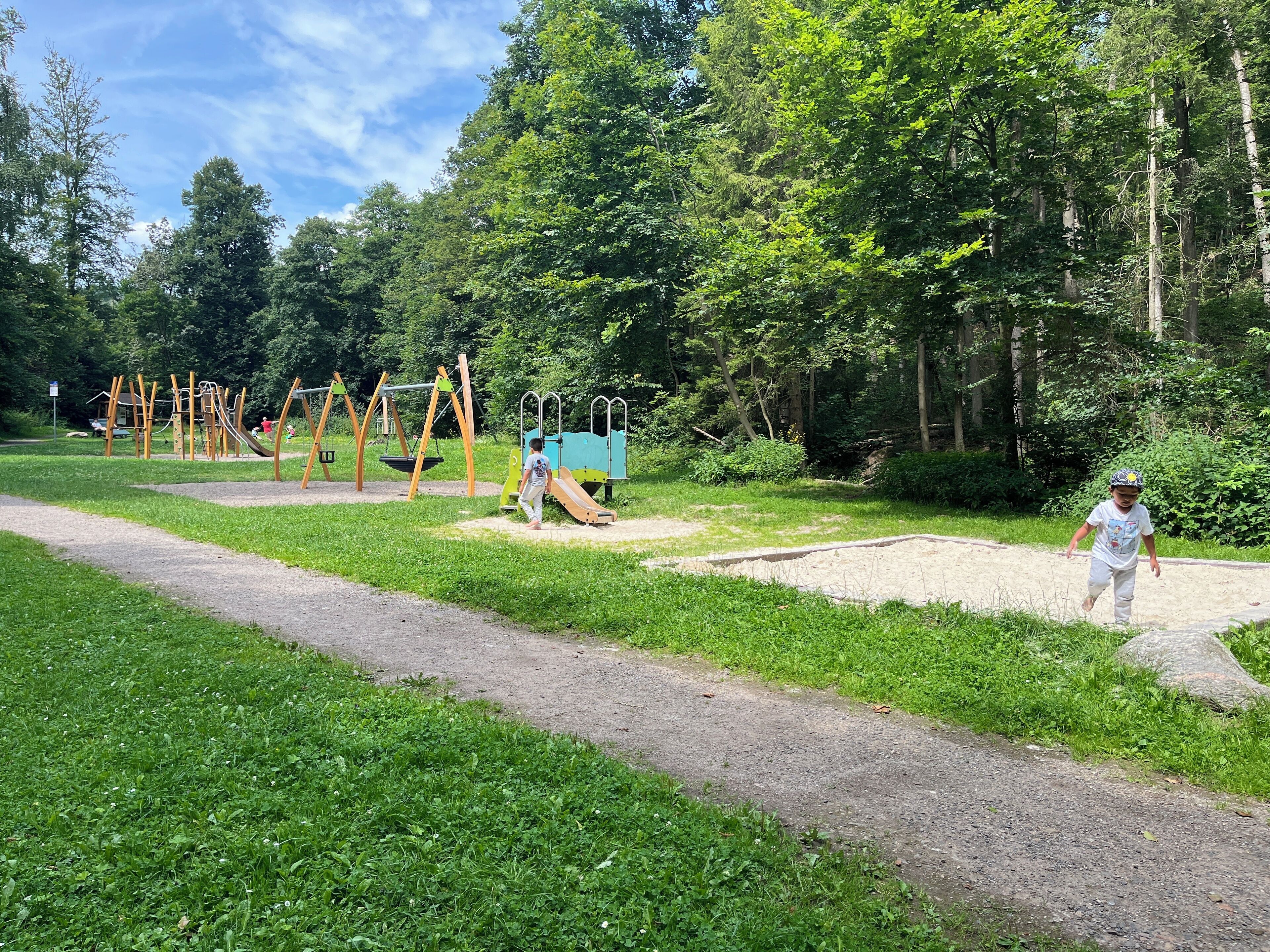 Un'area giochi con altalene e scivoli, circondata da alberi e un vialetto sterrato.