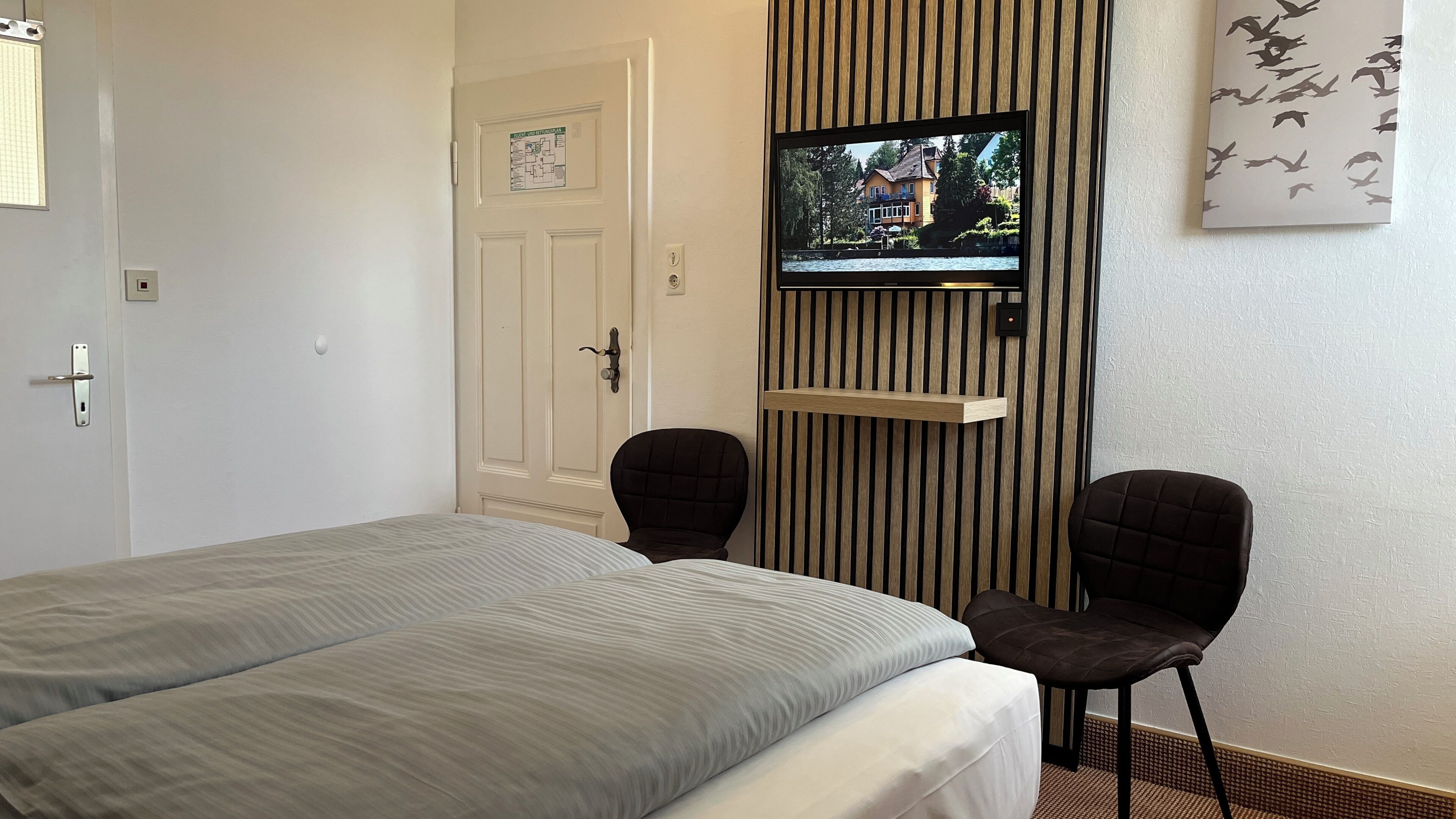 Ein Hotelzimmer mit einem Bett, zwei Sesseln, einem an einer Holzvertäfelung montierten Fernseher und einer Tür mit einer Karte.