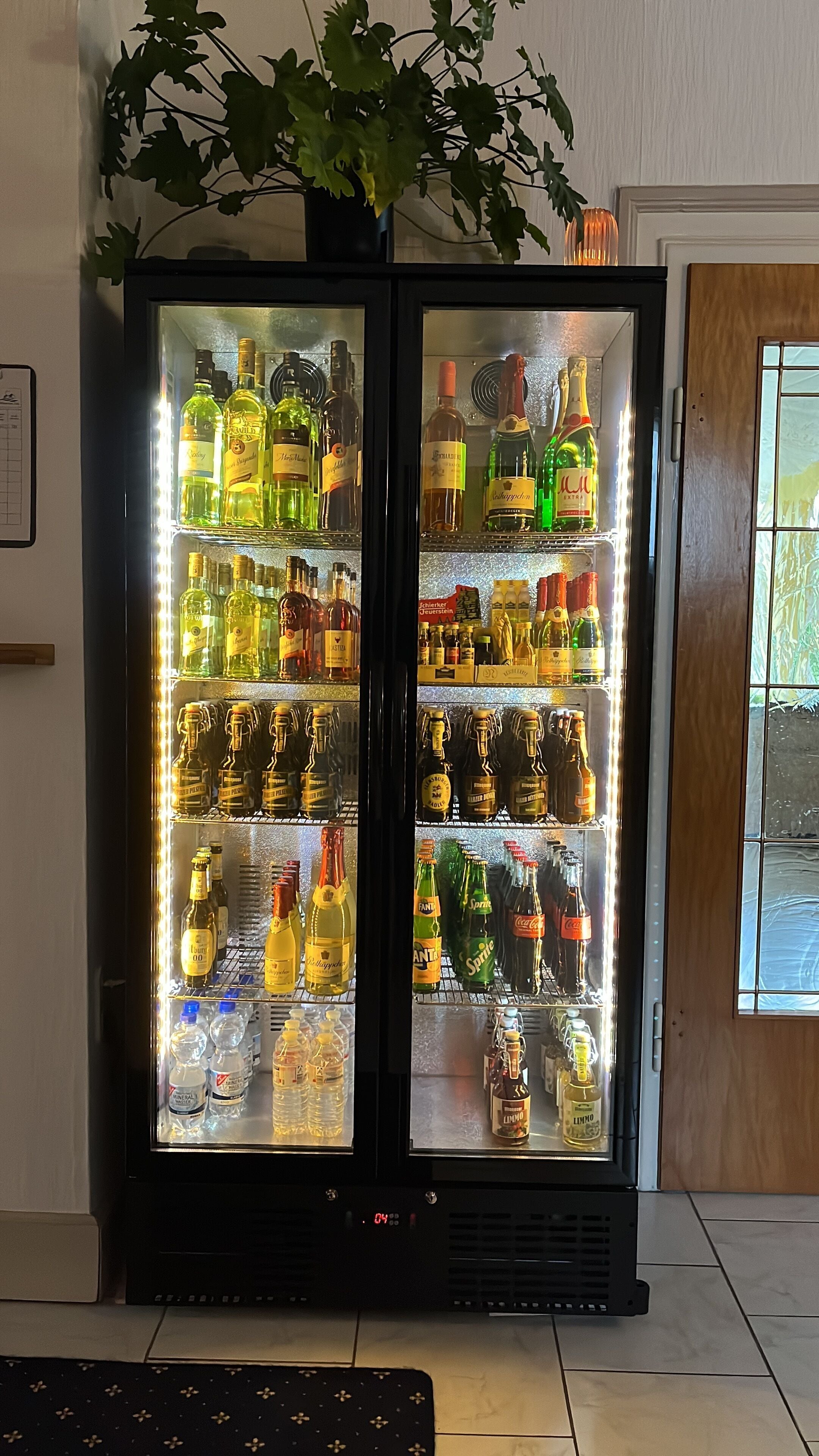 Ein Getränkekühlschrank, gefüllt mit verschiedenen Getränken, darunter Flaschen und Dosen, in einem Raum mit Holztür und Pflanze darauf.