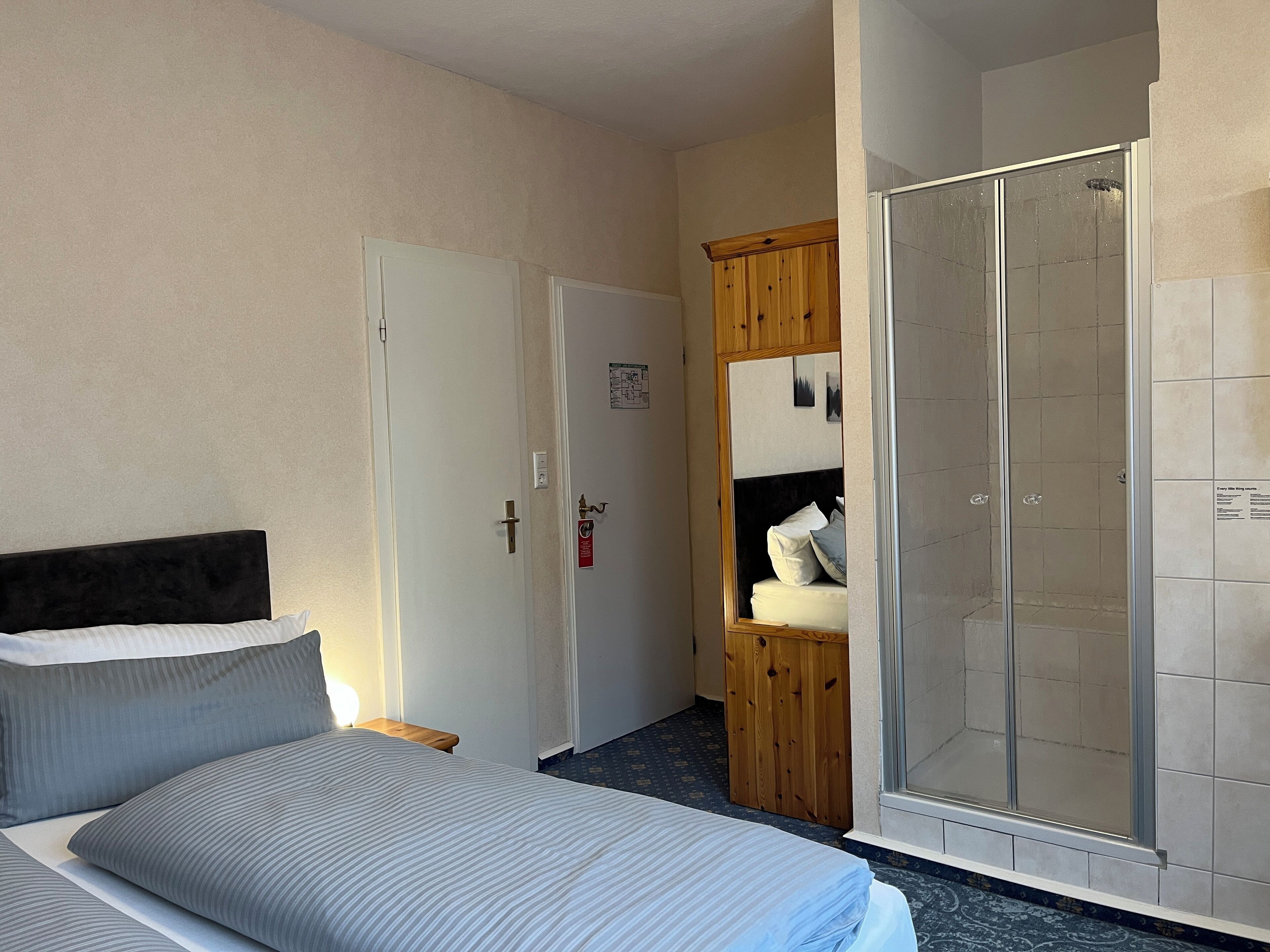 A hotel room with a bed, a wooden wardrobe, a shower area, and a door with a light switch.
