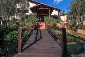 A wooden bridge leading to a building with a sloped roof and a view of a pond.