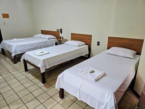 A hotel room with three single beds, each with a white bedspread and pillow, a wooden headboard, and a small bedside table with a lamp.