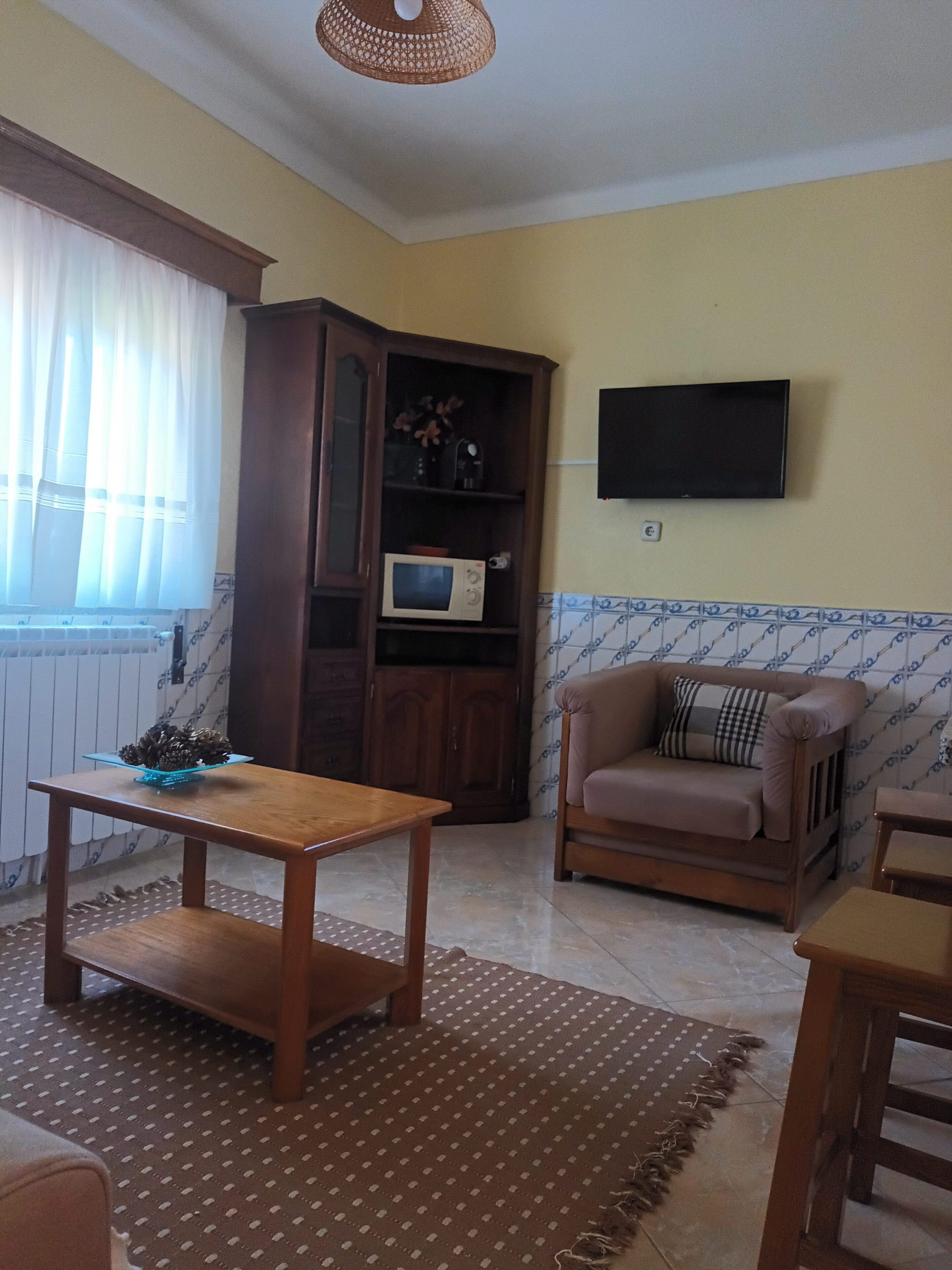 A living room with a wooden table, a sofa, a television mounted on the wall, and a cabinet with a microwave inside.