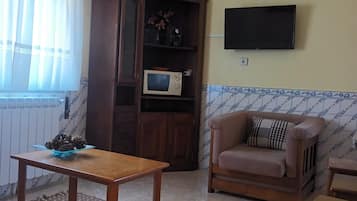 A living room with a wooden table, a sofa, a television mounted on the wall, and a cabinet with a microwave inside.