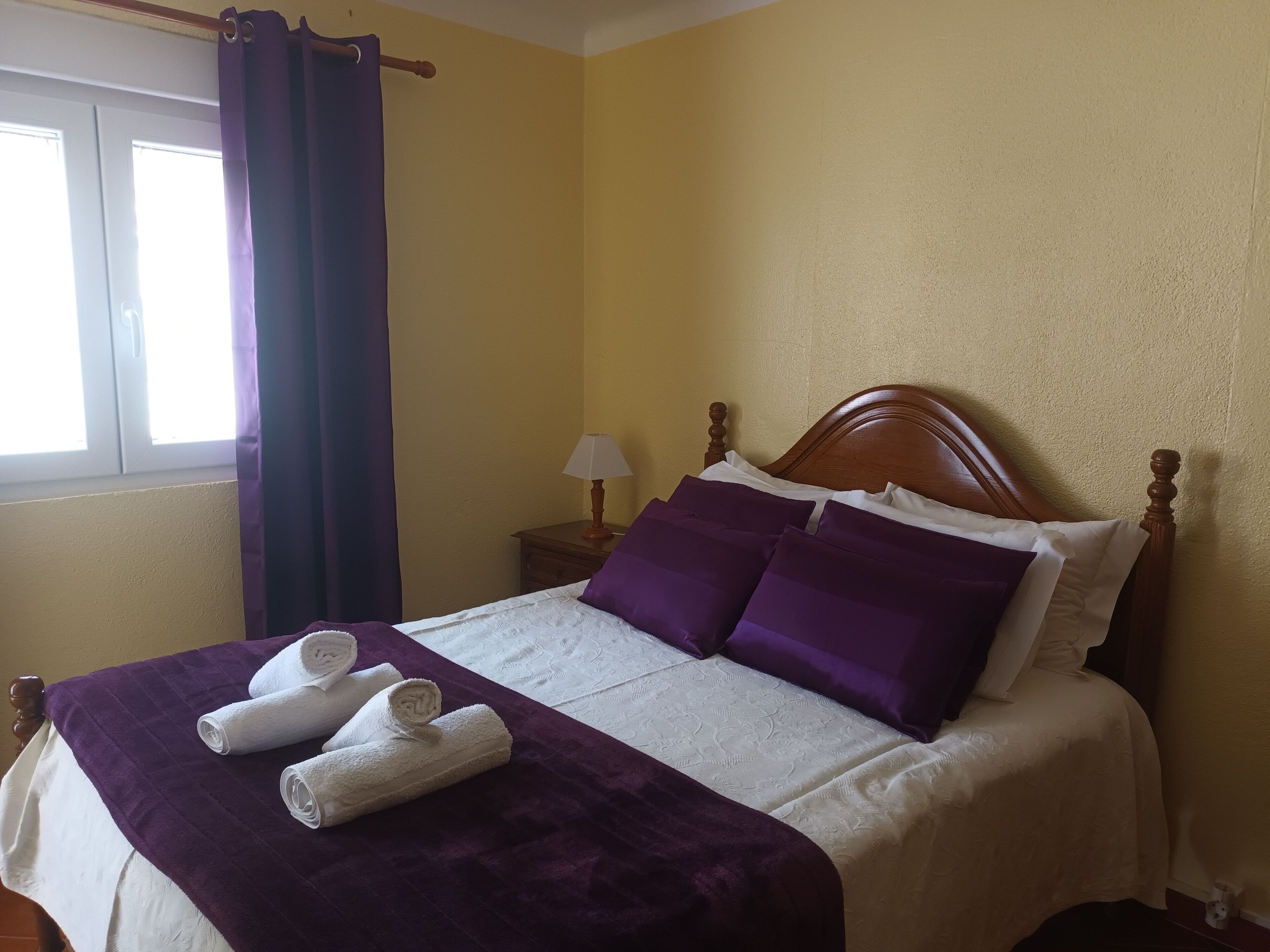 A neatly made bed with a purple blanket and pillows, a wooden headboard, and a nightstand with a lamp.