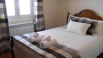 A neatly made bed with a checkered bedspread and pillows, a wooden headboard, and a window with curtains.