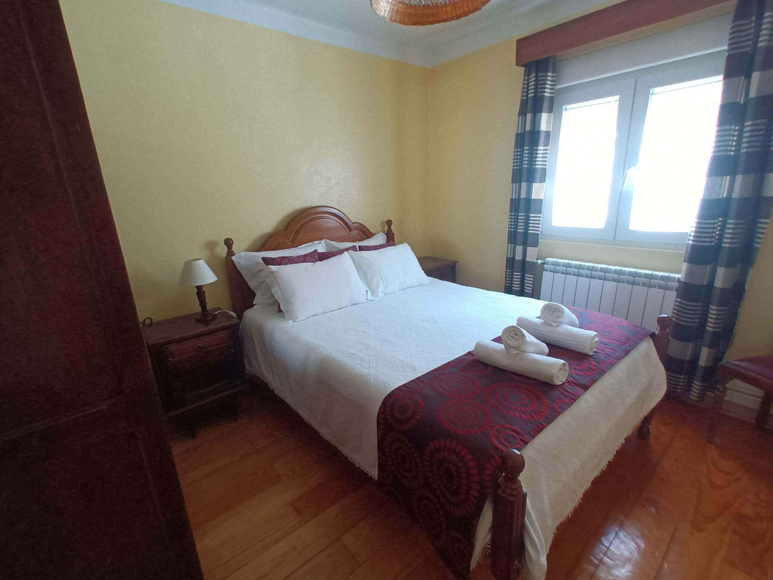 Una cama bien hecha con sábanas blancas y una colcha roja estampada, una mesita de noche de madera con una lámpara, y una ventana con cortinas.