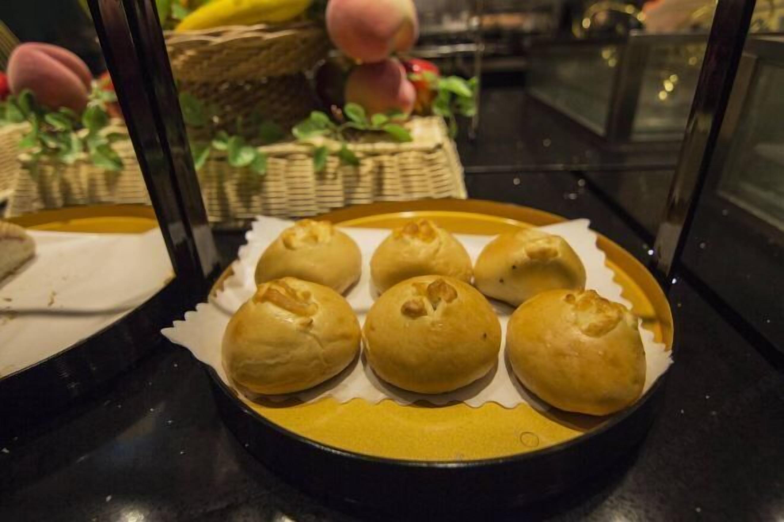 A tray of six golden-brown buns on a yellow plate, with baskets of fruits in the background.