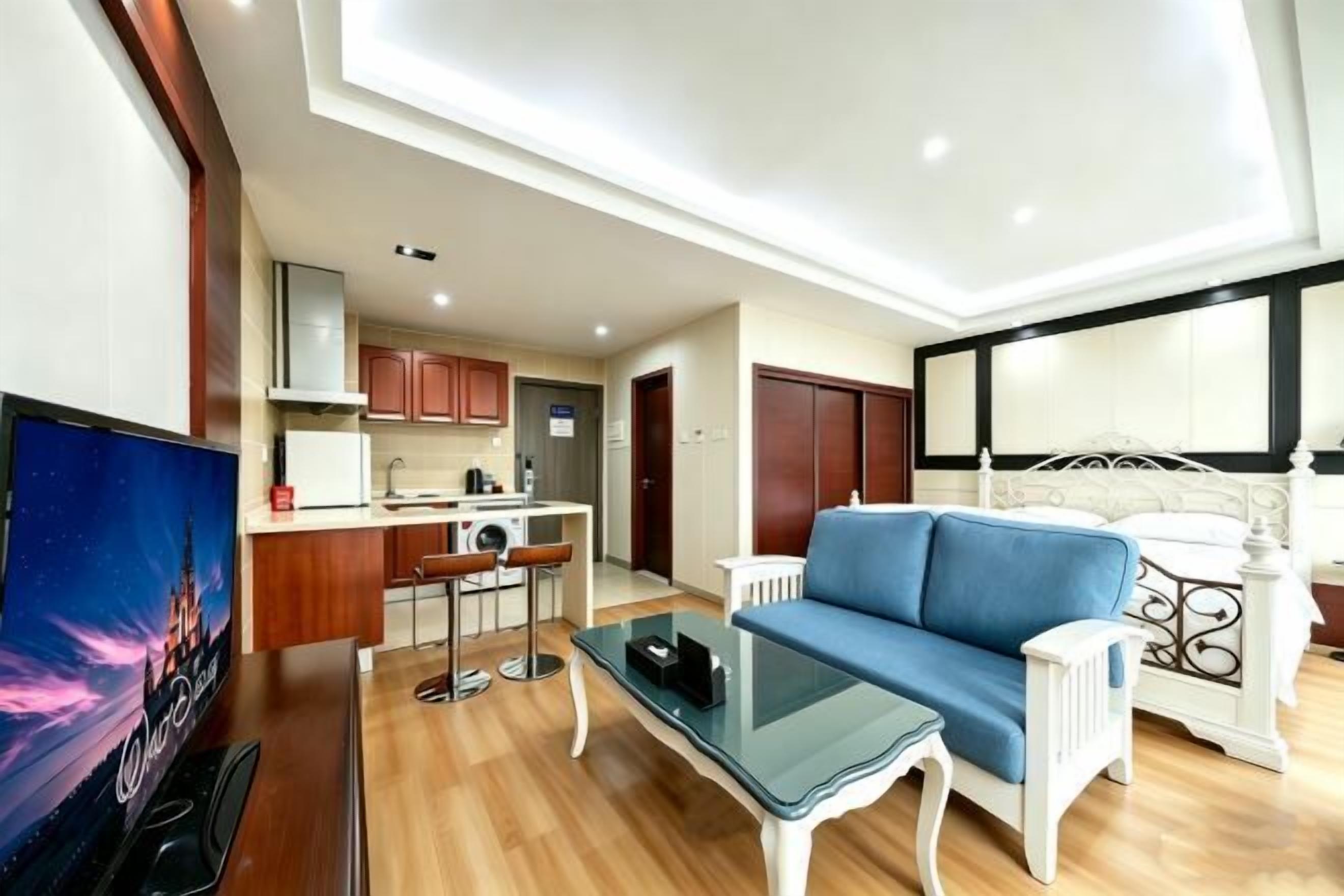 A modern living room with a sofa, a coffee table, and a TV. There is a kitchen area with a dining table and chairs. The room has a high ceiling with recessed lighting.