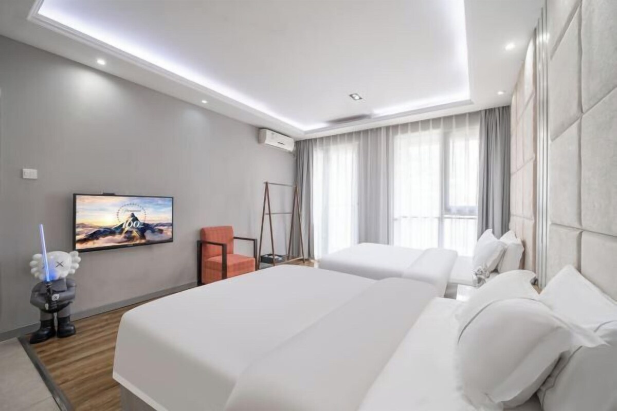 A hotel room with two beds, a flat-screen TV, a small sofa, and a large window with curtains.