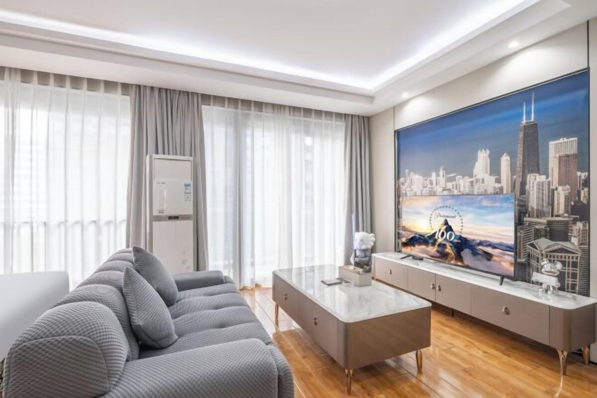 A modern hotel room with a sofa, a coffee table, a TV, and a large window with curtains.