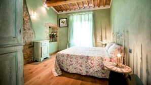 A bedroom with a wooden ceiling, a bed with floral bedding, a stone wall, and a small table with a candle. - Villa Meraviglia With Pool And Ac (Poggibonsi)