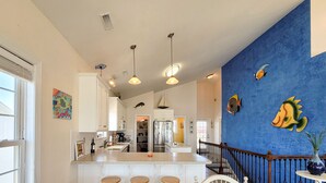 A spacious kitchen with a central island, white bar stools, pendant lighting, and a blue accent wall with fish decorations. - Dive into "Summer Days & Cooler Nights"! w/ pool! (Surf City)