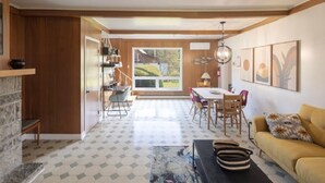 A modern living room with a stone fireplace, a yellow sofa, and a dining area with a view of the outdoors. - Maison Du Havre (Petit Duplex) (La Malbaie)
