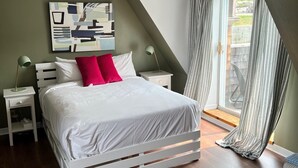 A bedroom with a white bed, a green accent wall, a modern abstract painting, and a view through a window with curtains. - Maison Du Havre (Petit Duplex) (La Malbaie)