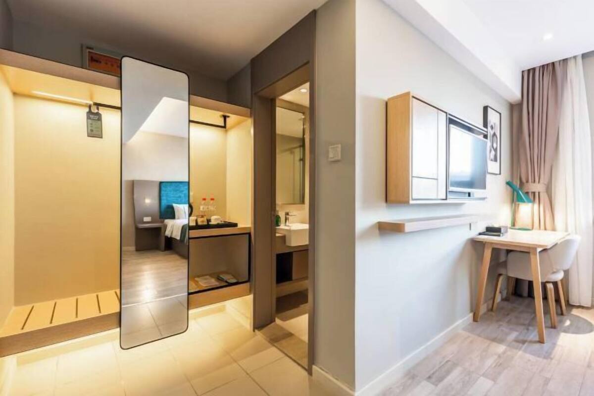 A modern hotel room with a large mirror, a desk with chairs, a television, and a bathroom with a sink and mirror.