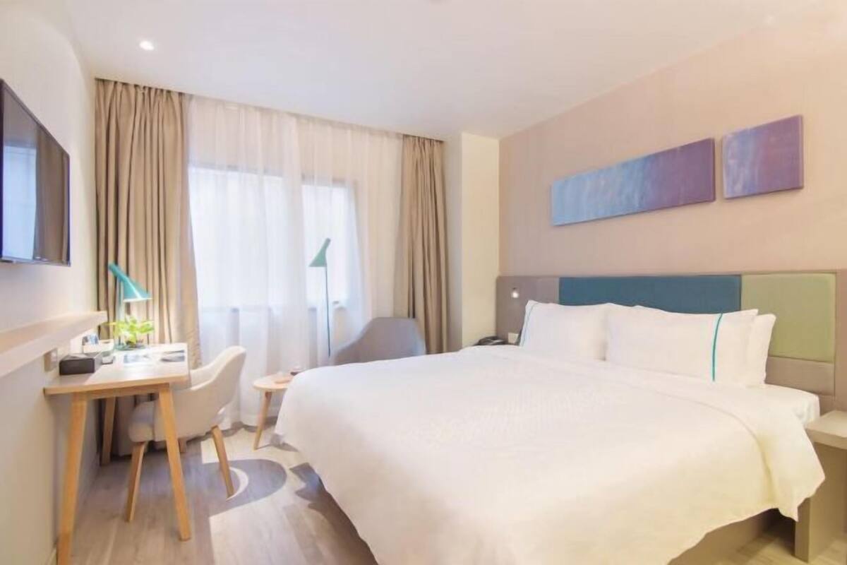 A hotel room with a bed, a desk with a chair, a TV, and two paintings on the wall.