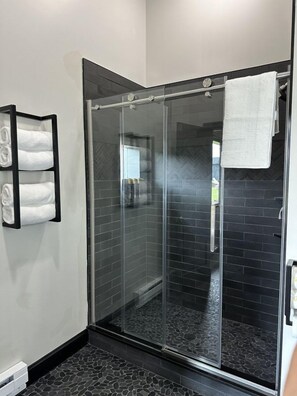 Une salle de bain moderne avec une douche à parois de verre, des murs recouverts de carreaux noirs et un porte-serviettes avec des serviettes pliées.