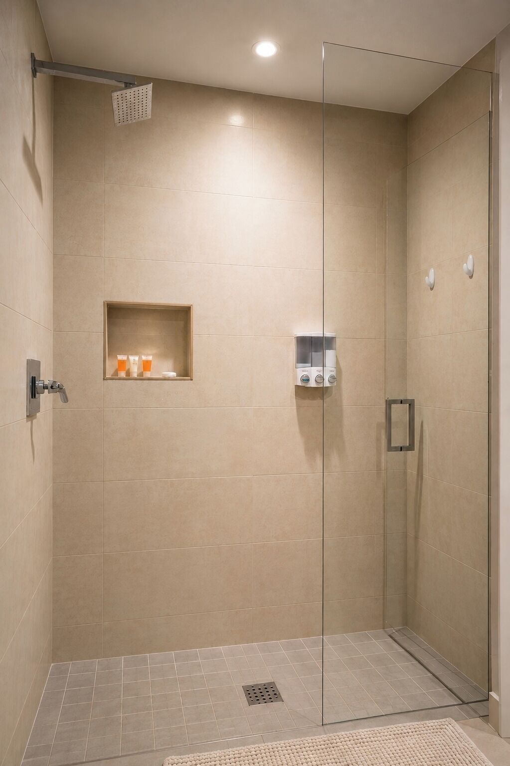 A glass-enclosed shower with a control panel, a shelf with toiletries, and a drain in the floor.