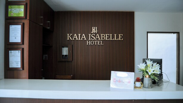 A reception desk at Kaia Isabelle Hotel with a wooden panel wall, framed documents, and a flower vase.