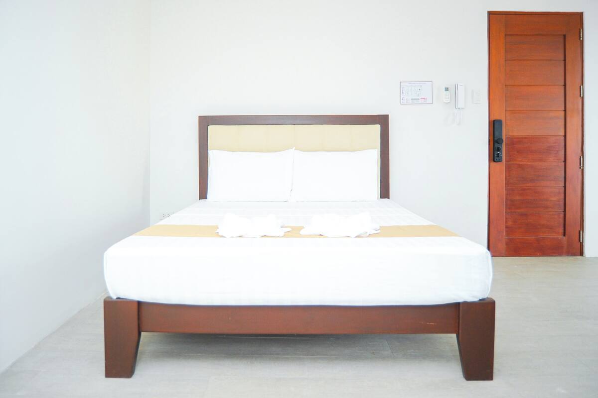 Ein ordentlich bezogenes Bett mit Holzrahmen und weißer Bettwäsche, eine Holztür rechts und eine schlichte weiße Wand mit Lichtschalter und Telefon.