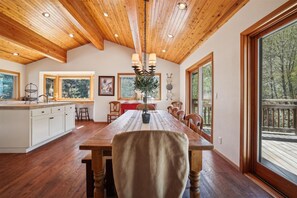 Un vaste espace dédié à la salle à manger, avec une table et des chaises en bois, une cuisine dotée de meubles blancs et de grandes fenêtres offrant une vue sur l’extérieur.