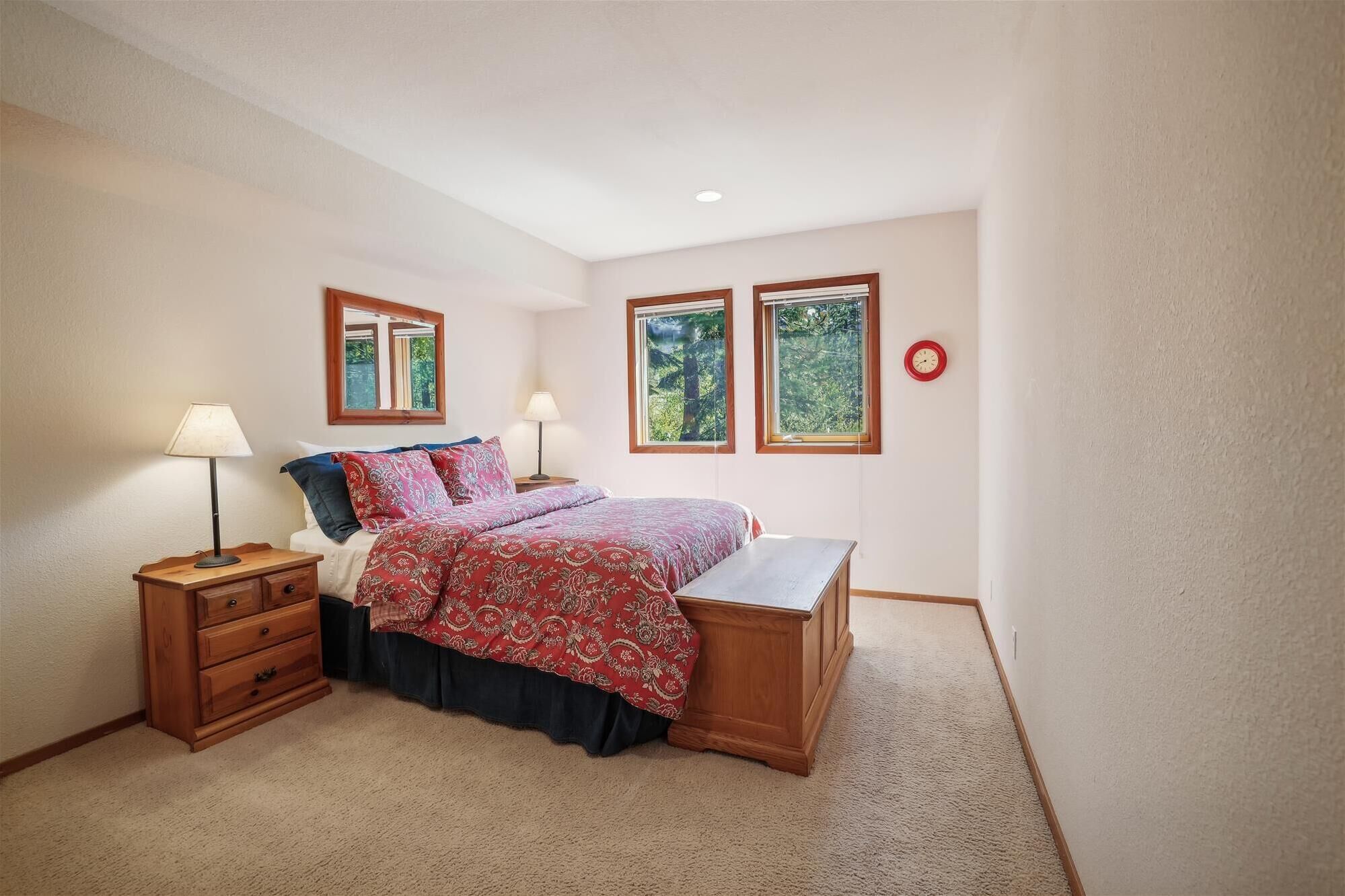 A bedroom with a bed, two bedside tables, a nightstand, a wooden bench, and two windows with views of greenery.