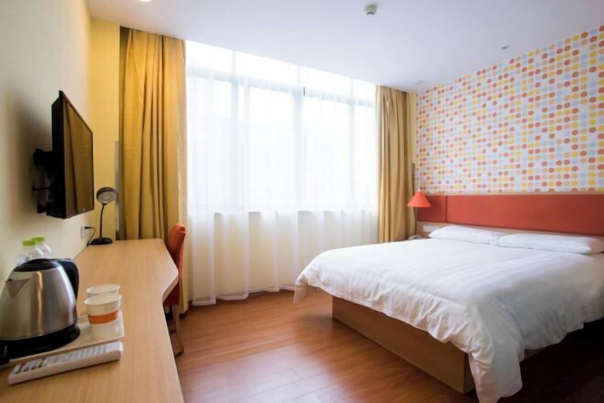A hotel room with a bed, a desk with a kettle, a cup, a remote control, and a TV.