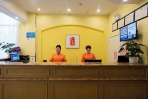 A reception desk with two staff members, a framed sign, a digital display, and a potted plant.