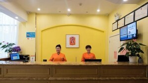 A reception desk with two staff members in orange uniforms, a yellow wall, a framed sign, and a computer monitor.