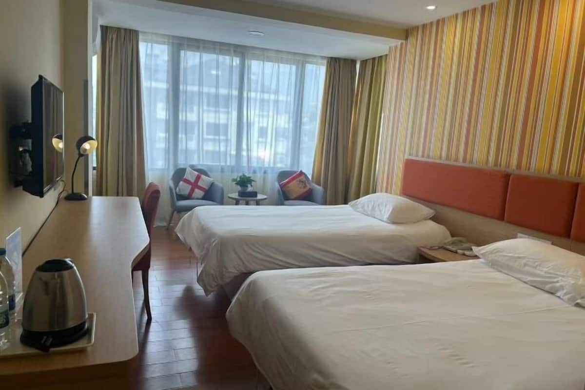 A hotel room with two beds, a desk with a kettle, a chair, a TV, and a window with curtains.