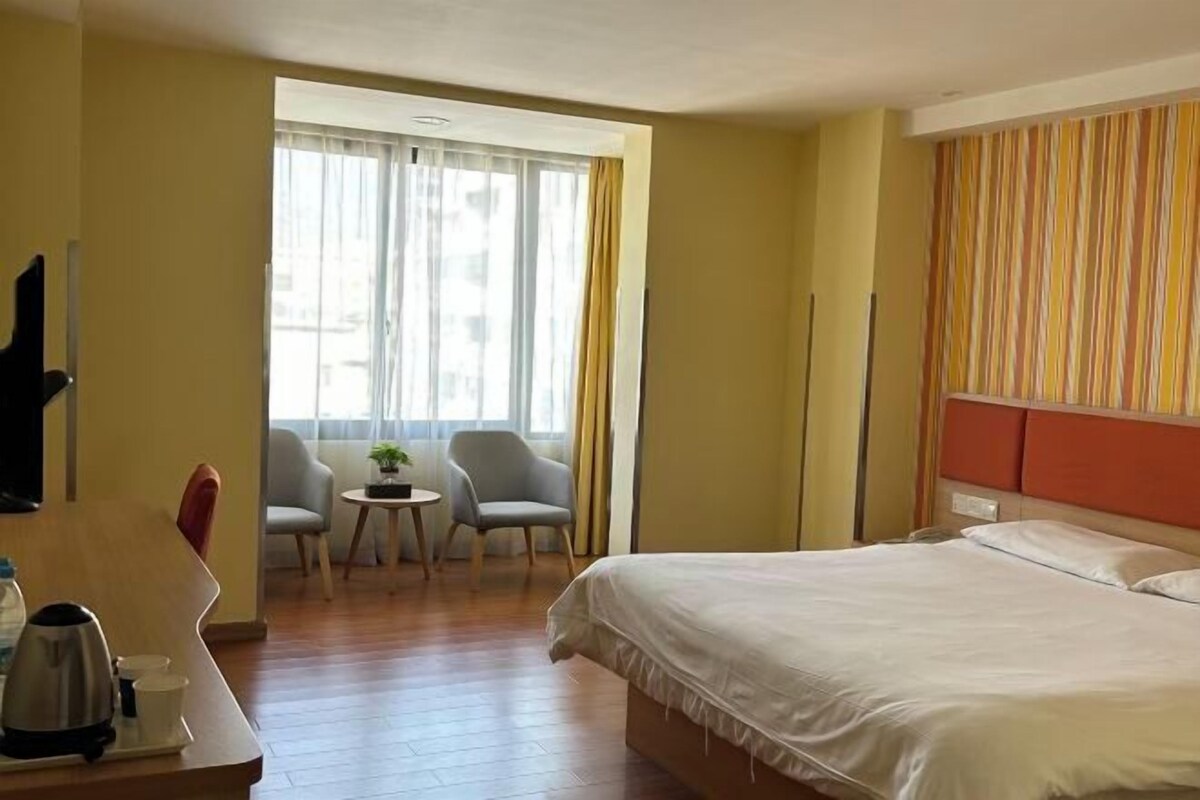 A hotel room with a bed, a desk with a kettle, two chairs, a small table with a plant, and a window with curtains.