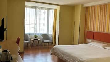 A hotel room with a bed, a desk with a kettle, two chairs, a small table with a plant, and a window with curtains.